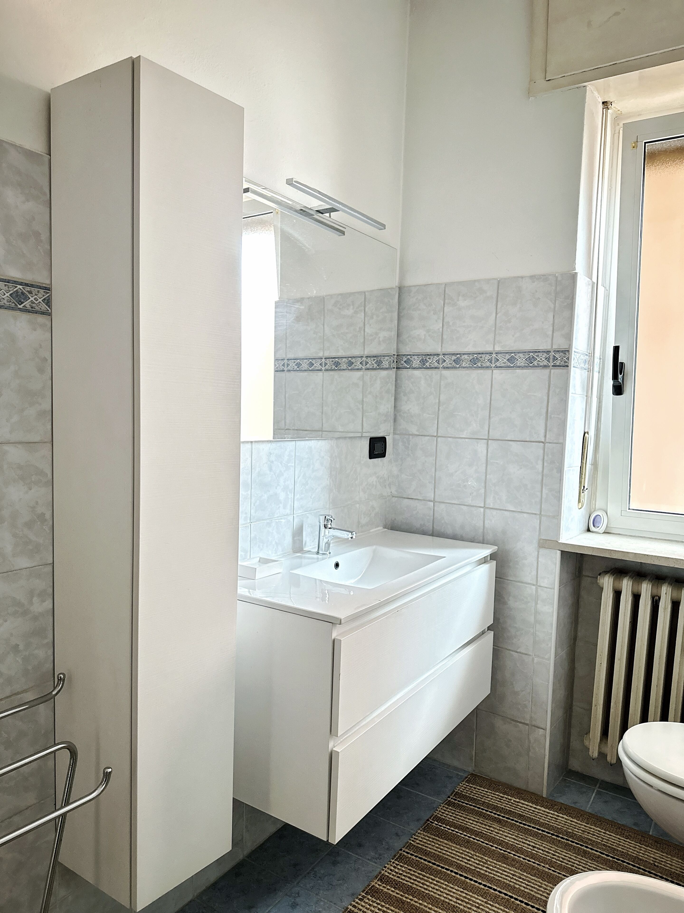 Een badkamer met een witte wastafel, spiegel en toilet. Er is een radiator onder het toilet en een handdoekhouder aan de linkerkant.