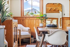 Un interior acogedor de madera con una mesa de comedor dispuesta para dos, una silla blanca y una ventana con vistas a montañas.