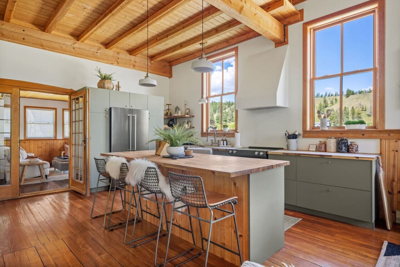 A spacious kitchen with wooden beams, a large island with bar stools, and a view of a green landscape through large windows.