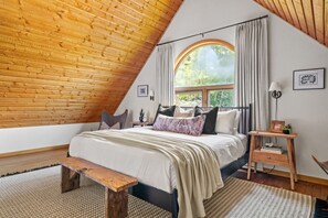 A bedroom with a sloped ceiling, a large window with curtains, a bed with multiple pillows, a bedside table with a lamp, and a small plant. - Smith`s Church House | 1903 | Fireplace | Iconic (Wilmer)
