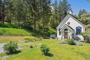 A small white house with a gabled roof, surrounded by a lush green landscape and tall trees. - Smith`s Church House | 1903 | Fireplace | Iconic (Wilmer)