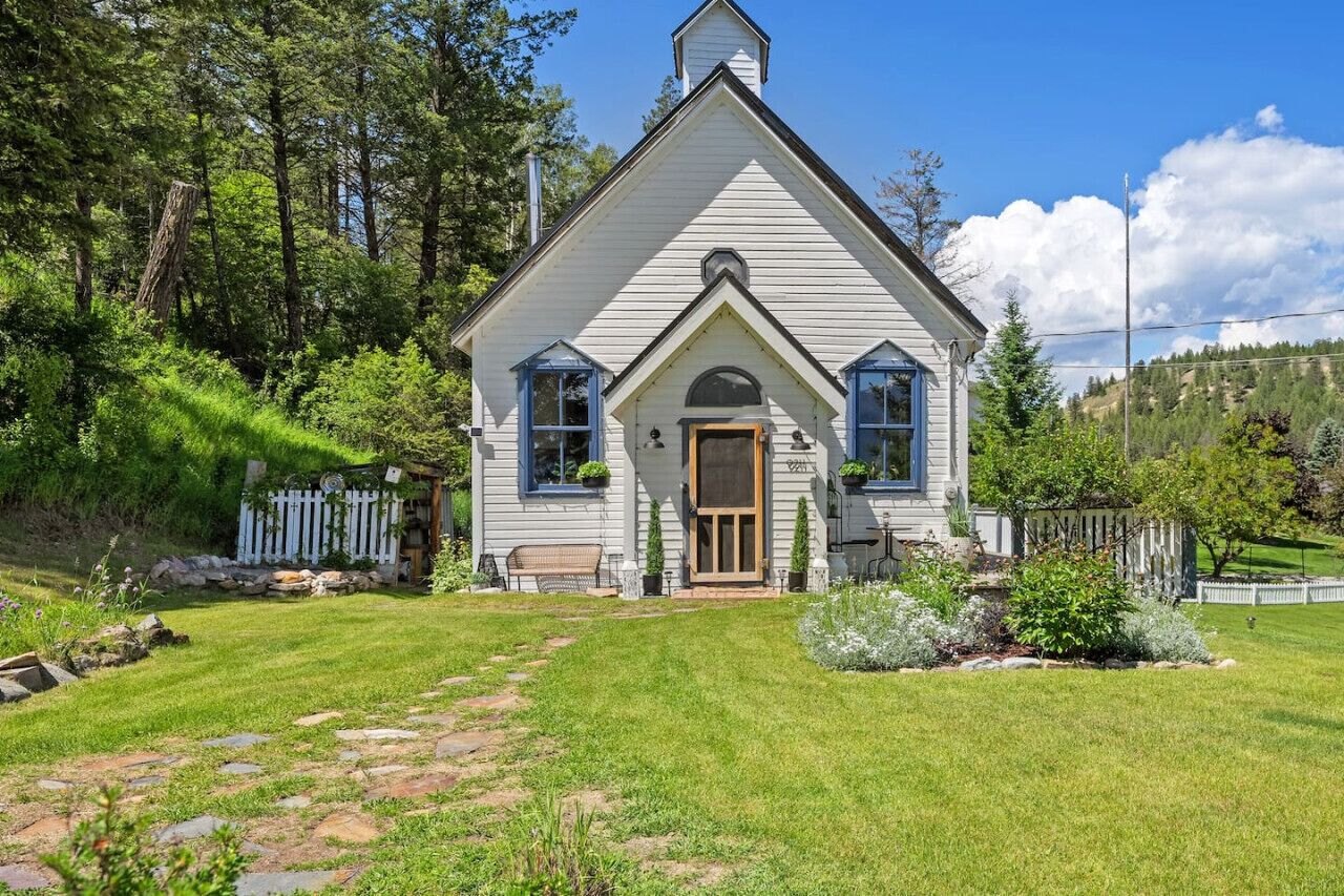 Une maison blanche, de plain-pied, avec un toit à pignon, un porche à l’avant et un petit jardin.