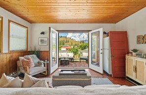 A cozy living room with a wooden ceiling, a sofa, a coffee table, and a view of a balcony with outdoor furniture. - Smith`s Church House | 1903 | Fireplace | Iconic (Wilmer)