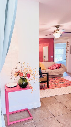 Una sala de estar con un escritorio rosa, un florero con flores, un ventilador de techo, un sofá color beige y una alfombra estampada.