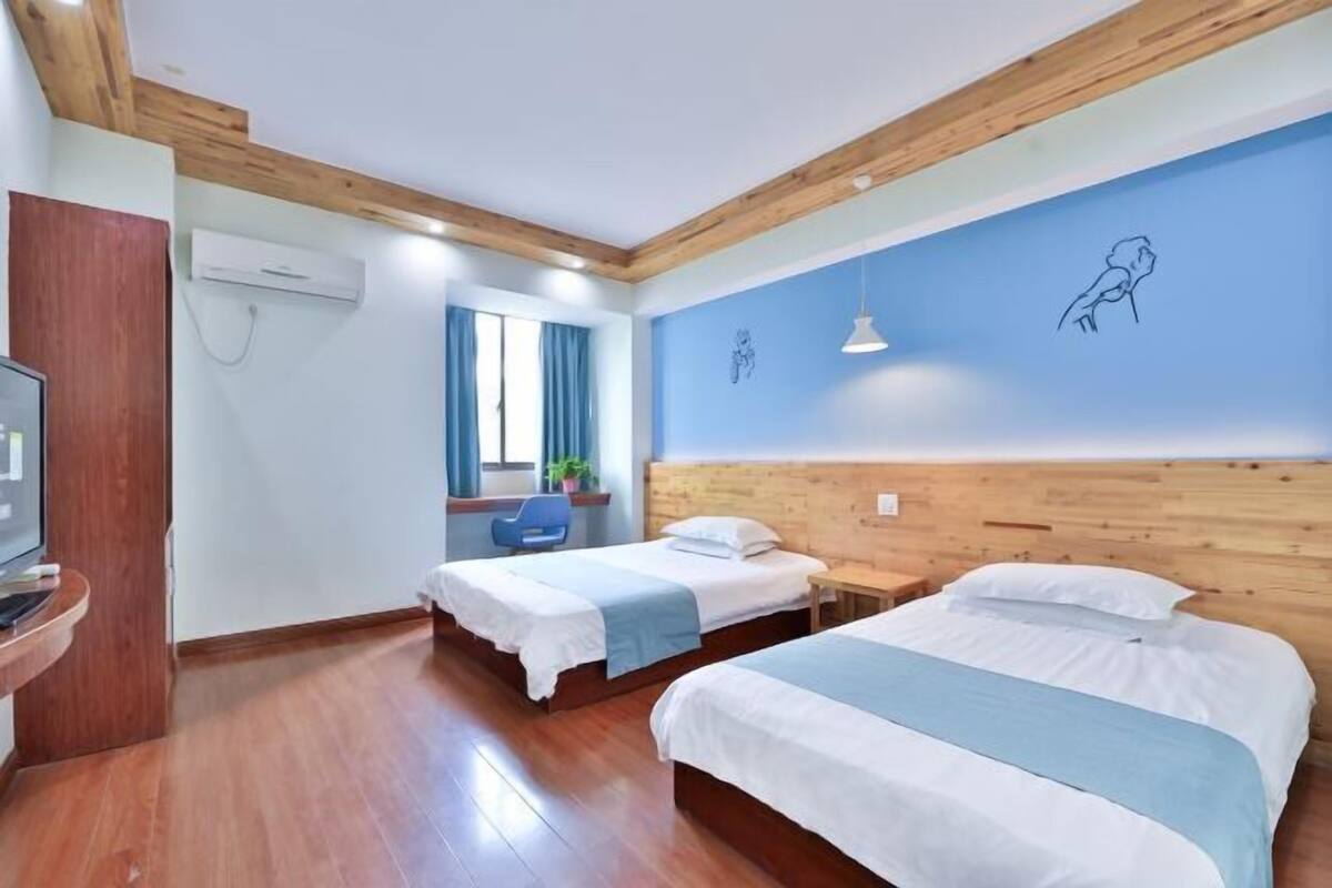 Ein Hotelzimmer mit zwei Betten, Holzbalkendecke, blauer Wand mit Vogelgemälde, Fenster mit Vorhängen und Schreibtisch mit Stuhl.