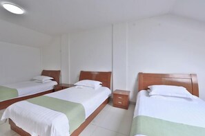 A hotel room with three single beds, each with a wooden headboard and white bedding. There is a small wooden nightstand between the beds.