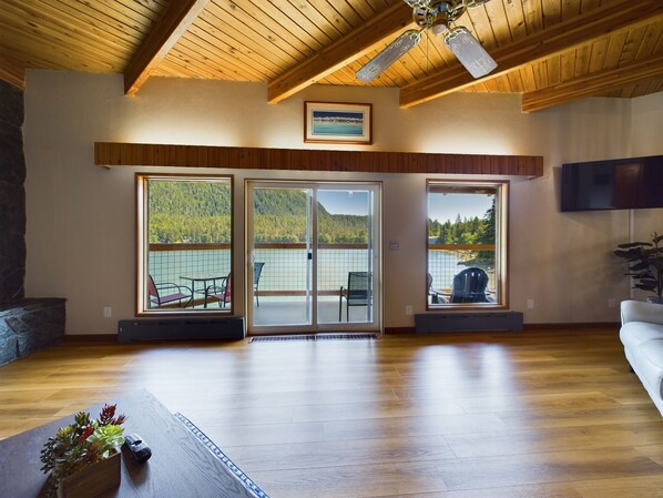 A spacious living room with wooden flooring, a ceiling fan, and large sliding glass doors leading to a balcony with a view of a lake and forest.