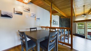 A dining area with a wooden table and chairs, framed artwork on the walls, and a view of the outdoors through large windows. - Stunning Oceanfront Home w/ Mountain Views! (Juneau)