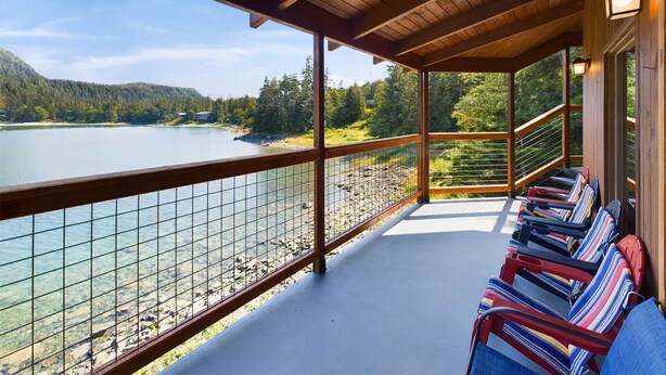 A wooden deck with a railing overlooking a lake, with several striped chairs and a view of the surrounding forest. - Stunning Oceanfront Home w/ Mountain Views! (Juneau)