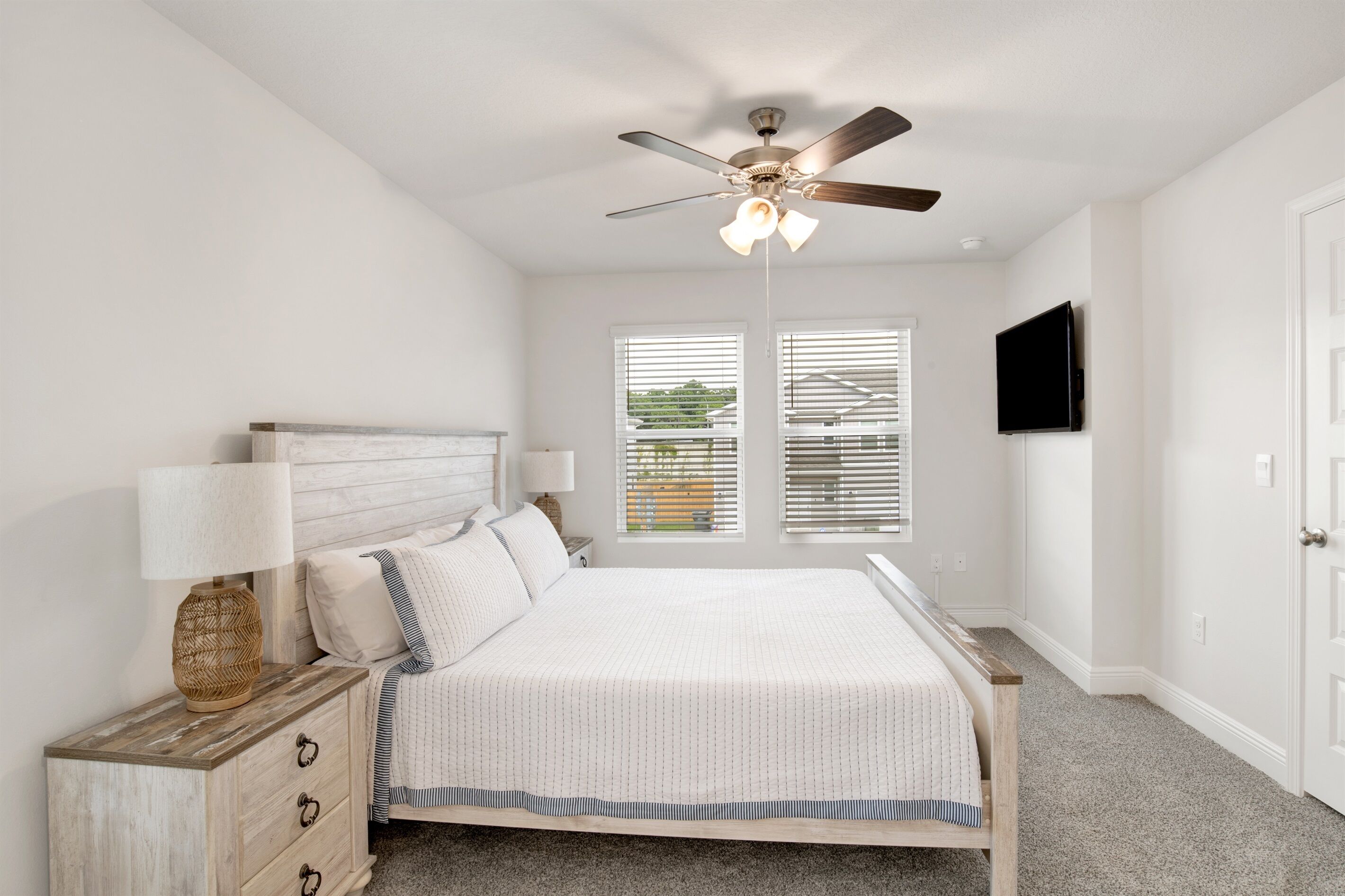 A bedroom with a bed, a nightstand, a lamp, a ceiling fan, a TV, and two windows with blinds.