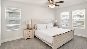Quarto com cama de madeira, paredes brancas e ventilador de teto. Há duas mesas de cabeceira com luminárias e um criado-mudo com abajur.