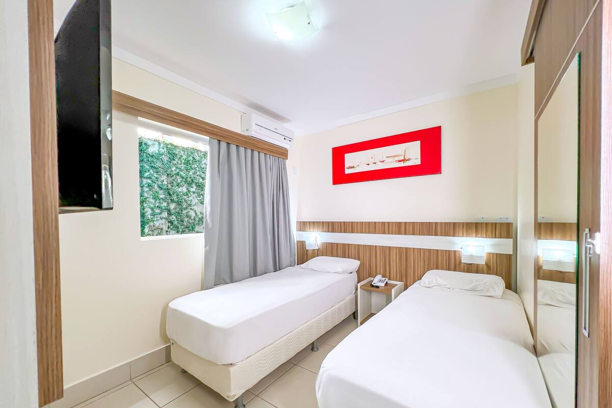 Une chambre d’hôtel avec deux lits simples, une fenêtre avec des rideaux, un climatiseur fixé au mur et un tableau encadré au-dessus des lits.