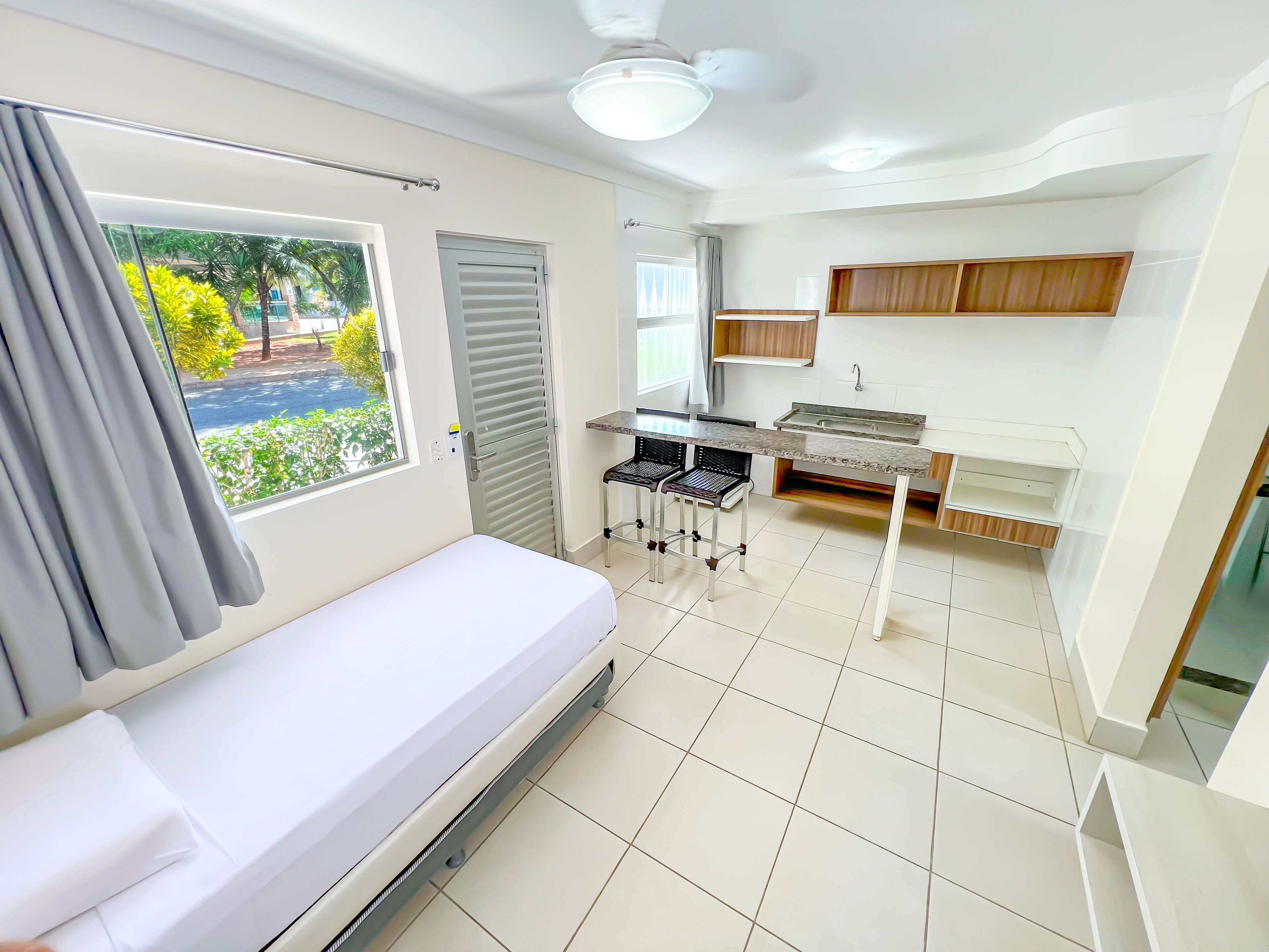 A compact room with a bed, a small kitchenette, a window with a view of greenery, and a ceiling fan.