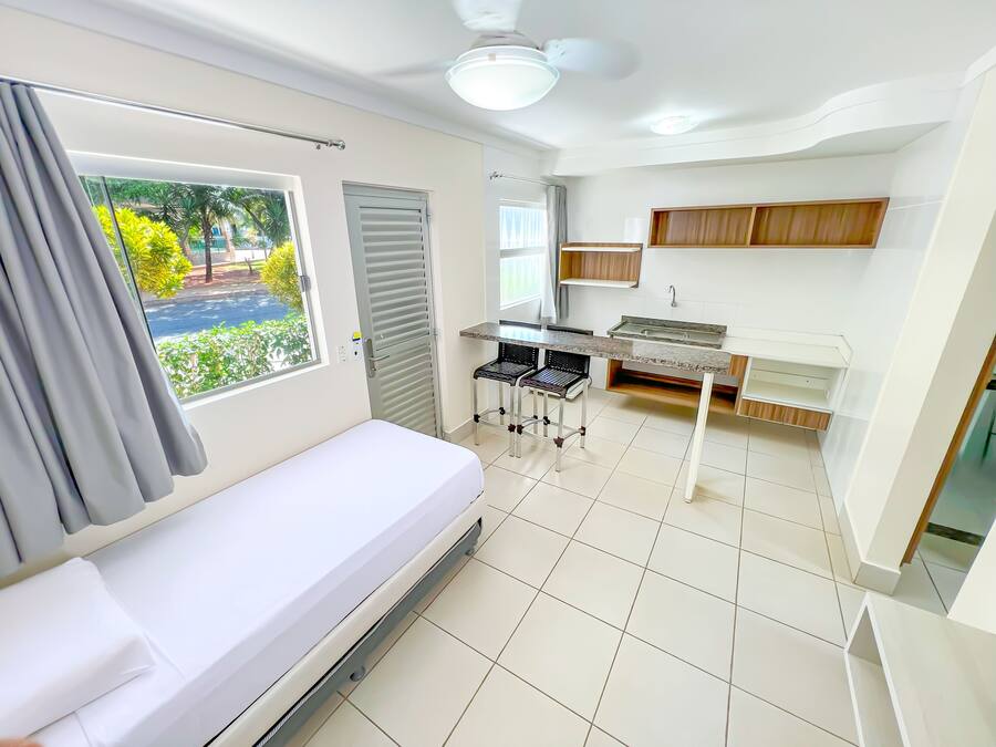 A compact room with a bed, a small kitchenette, a window with a view of greenery, and a ceiling fan.