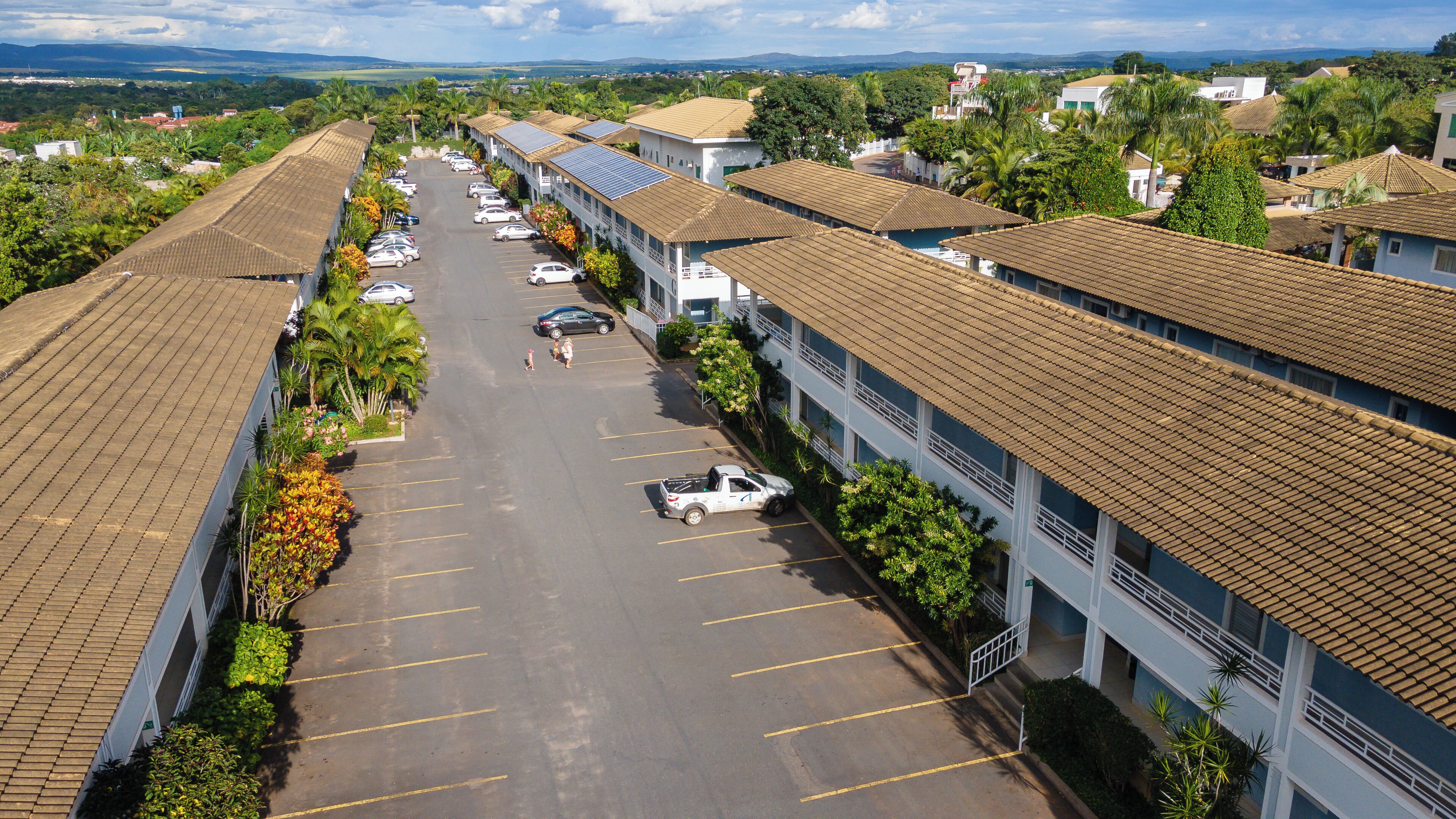 A parking lot with several cars, a white pickup truck, and a row of buildings with tiled roofs and balconies.