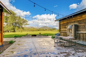 A patio with a picnic table and string lights, a wooden fence, and a view of a grassy field and mountains. - Spacious Afton Home: Patio, Grill & Mountain Views (Afton)