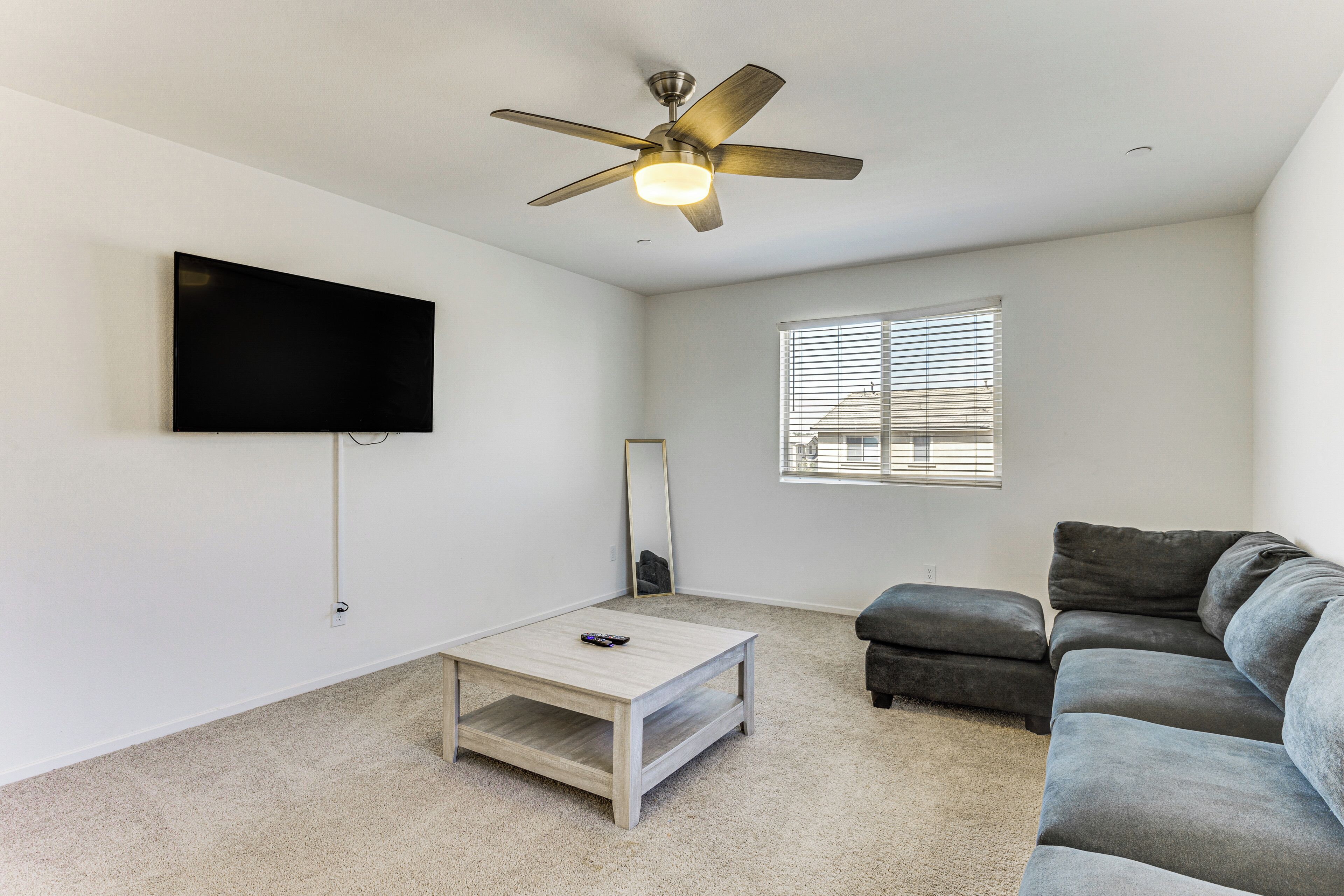 A living room with a ceiling fan, a flat-screen TV, a coffee table, a gray sofa, and a window with blinds.