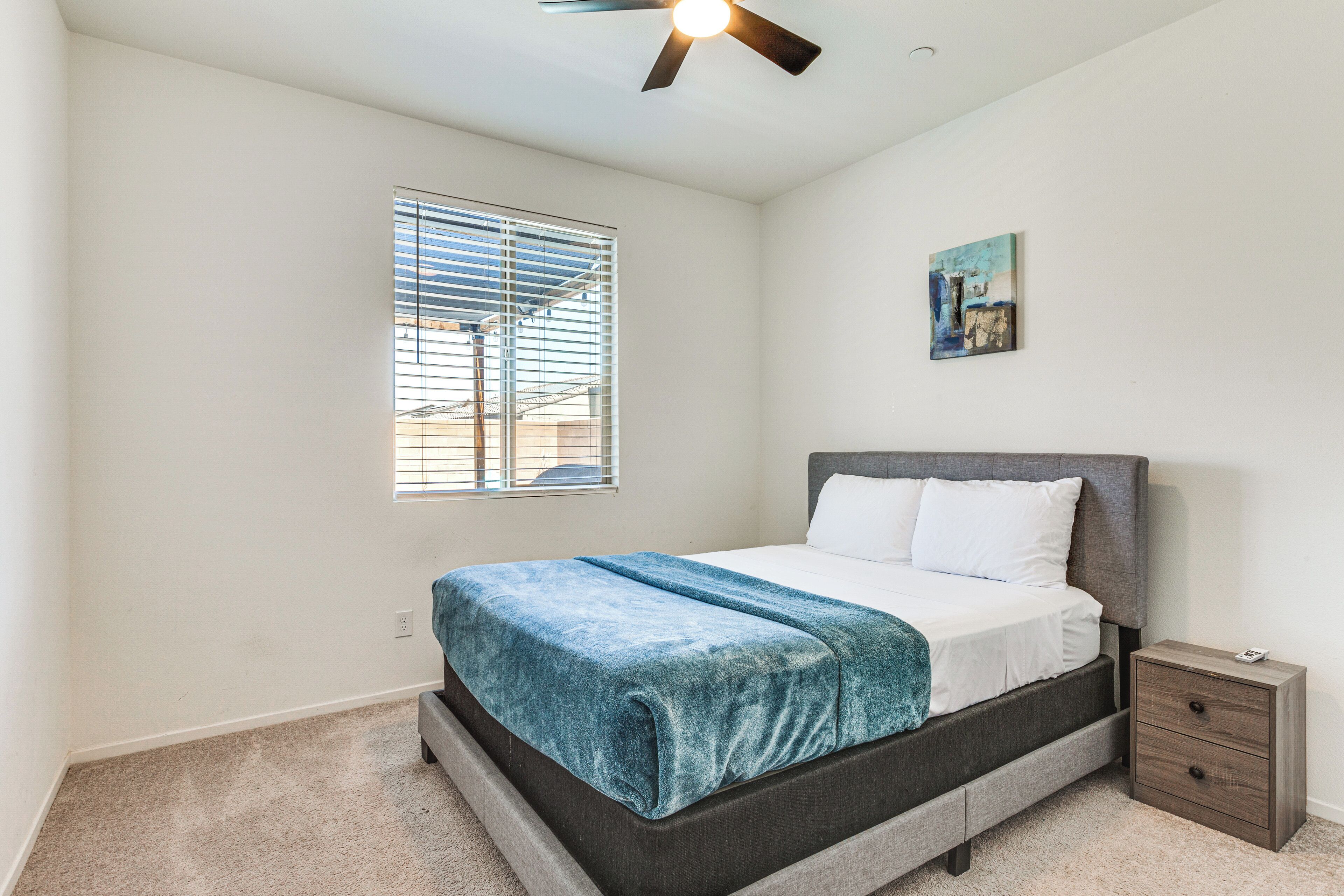 A bedroom with a bed, a nightstand, a ceiling fan, a window with blinds, and a wall-mounted artwork.