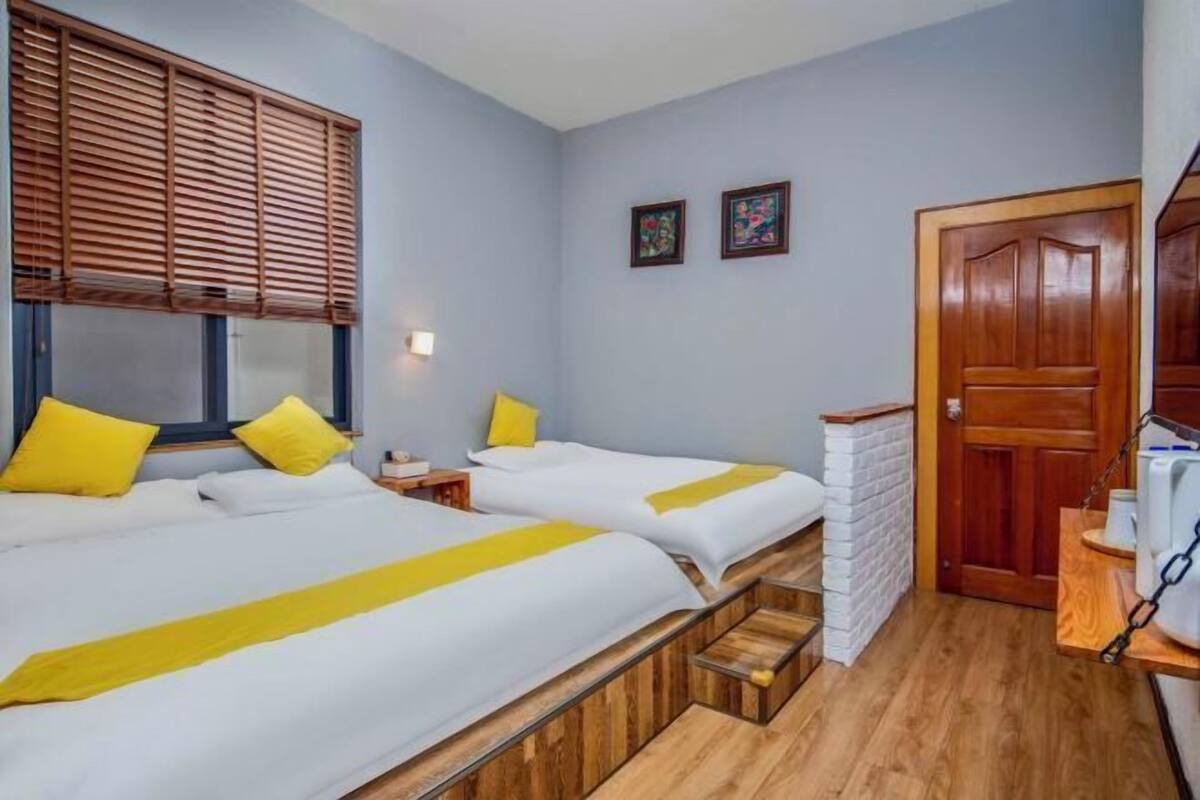 A hotel room with two beds, wooden flooring, a brick wall, a wooden door, and a small table with a kettle.