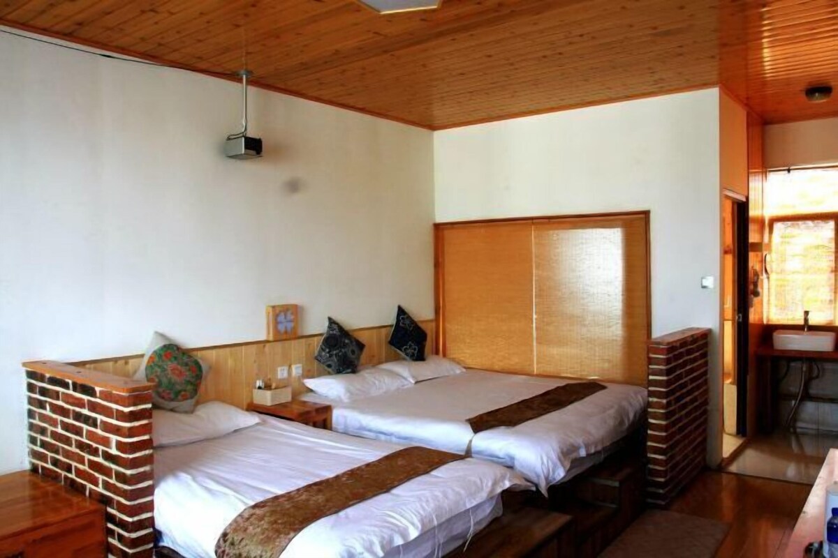 Ein Zimmer mit zwei Betten, einem Holz-Kopfende, einer Ziegelwand als Blickfang, einem kleinen Nachttisch und einem Fenster mit Tageslicht.