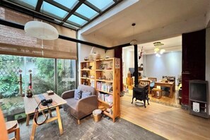 A spacious living area with a large window, a wooden table, a sofa, and a bookshelf. - Buxianshan Guesthouse (Dali City)
