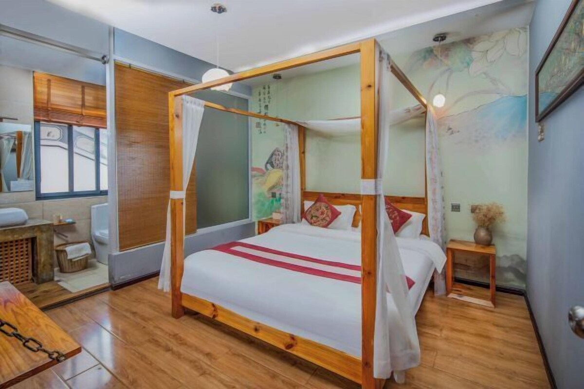 Ein Bett mit Baldachin, dessen Gestell aus Holz gefertigt ist und mit weißer Bettwäsche bezogen ist, ein kleiner Nachttisch und ein an der Wand befestigtes Regal.