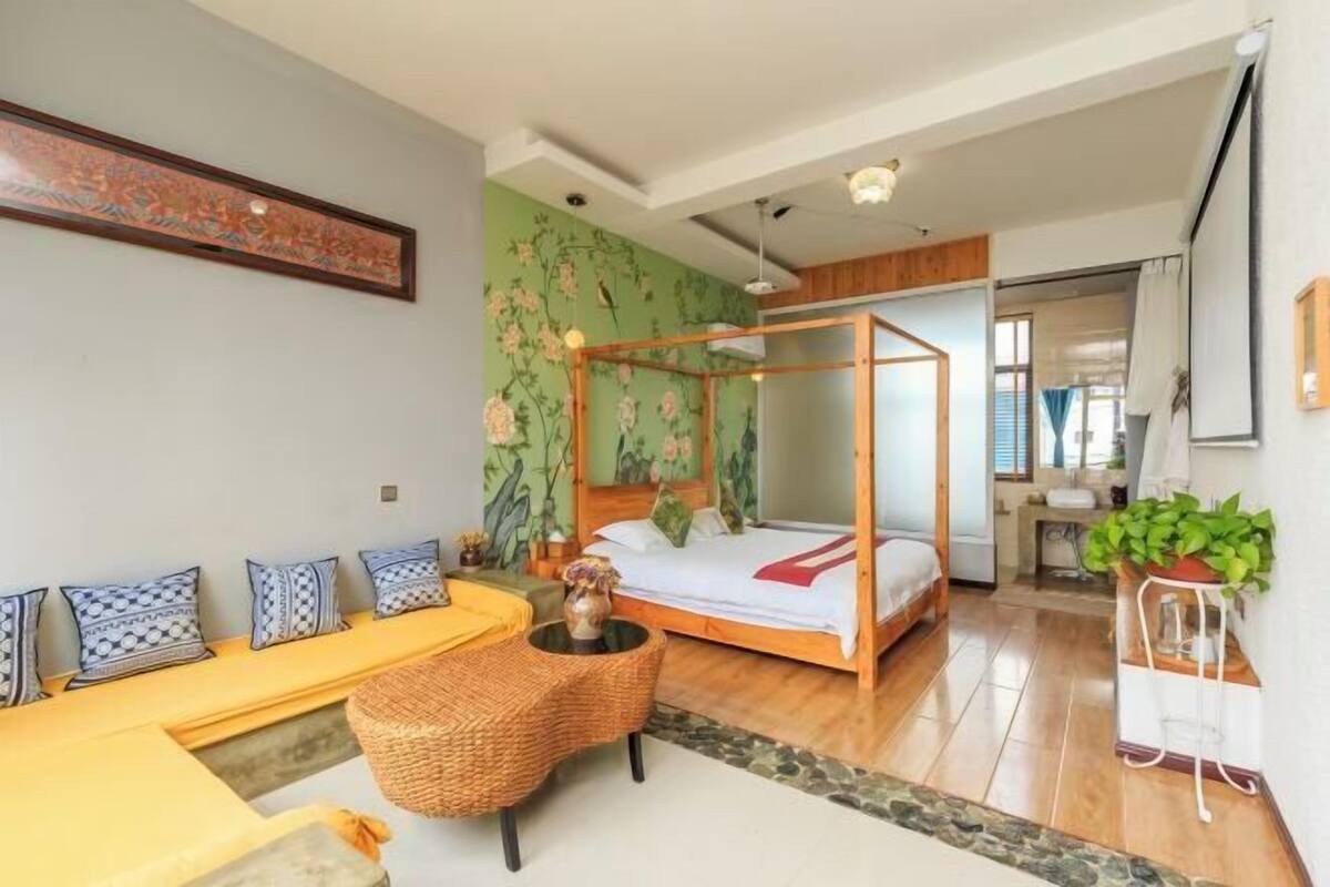A room with a four-poster bed, a sofa with patterned cushions, a small table with a vase, and a potted plant.