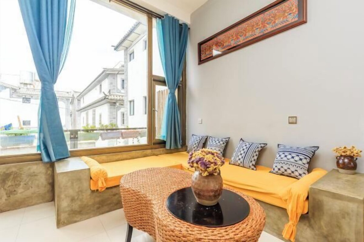 Ein Zimmer mit einer Sofagarnitur, einem runden Tisch mit einer Vase und einem Fenster mit Vorhängen.