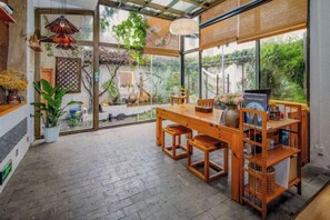 A dining area with a wooden table and chairs, a hanging light fixture, and a view of greenery through large windows. - Buxianshan Guesthouse (Dali City)