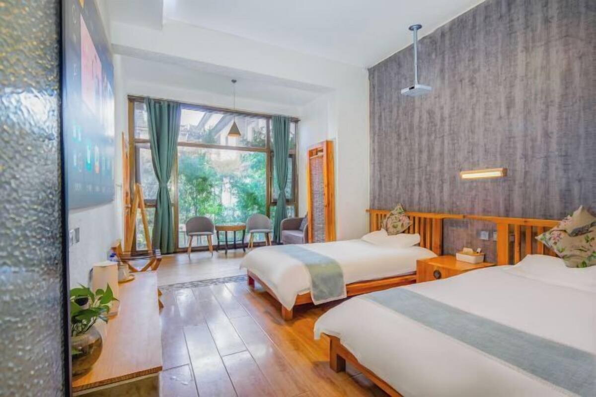 A hotel room with two beds, a wooden floor, a large window with curtains, a ceiling light, and a wall-mounted shelf.