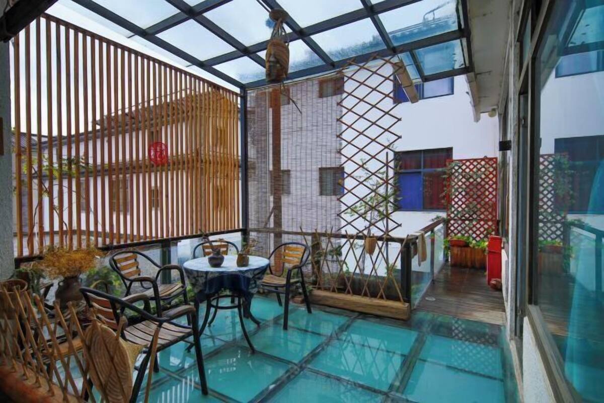 A covered outdoor patio with a glass table and chairs, a wooden fence, and a view of a building in the background.