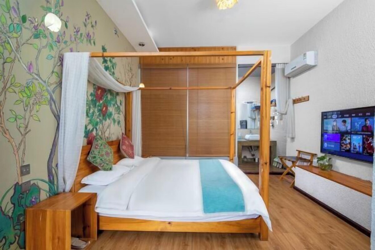 Ein Zimmer mit einem hölzernen Himmelbett, einem an der Wand befestigten Fernseher und einem durch eine Glastür einsehbaren Badezimmer.