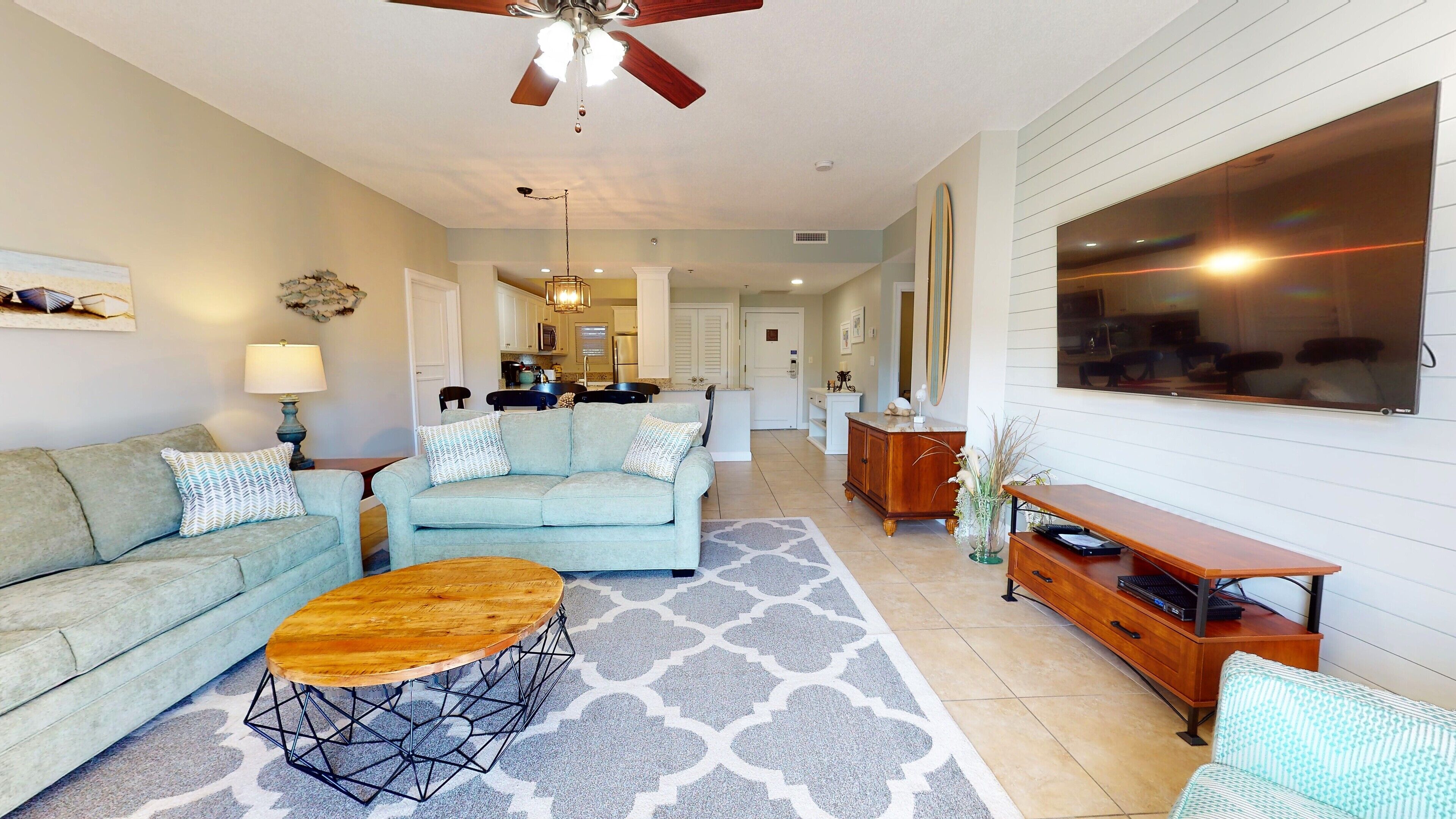 A living room with a sofa, coffee table, and a dining area with a table and chairs. There is a kitchen in the background.