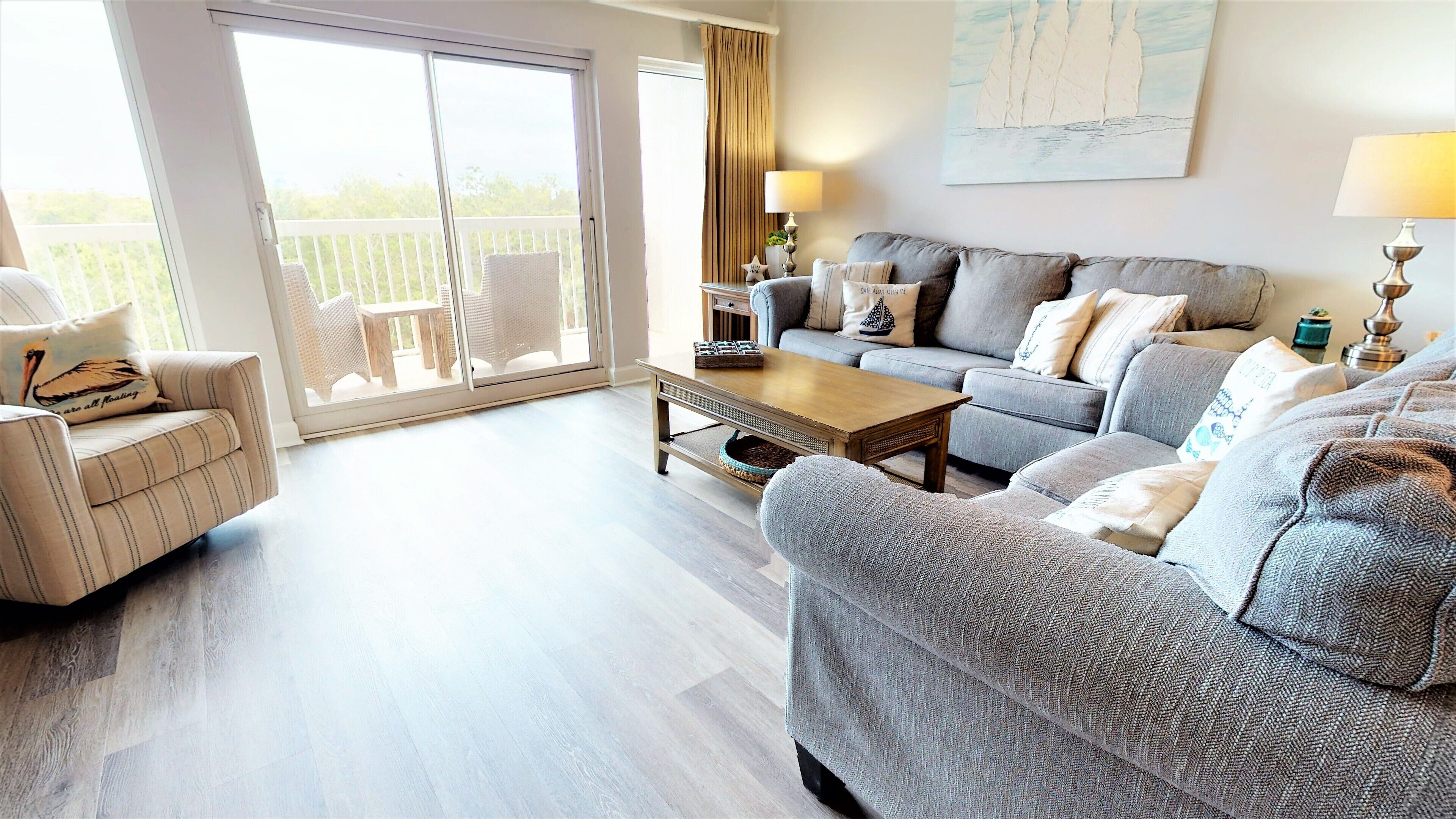 A spacious living room with a sofa, armchair, and coffee table. There is a sliding glass door leading to a balcony with outdoor furniture. A painting is visible on the wall.