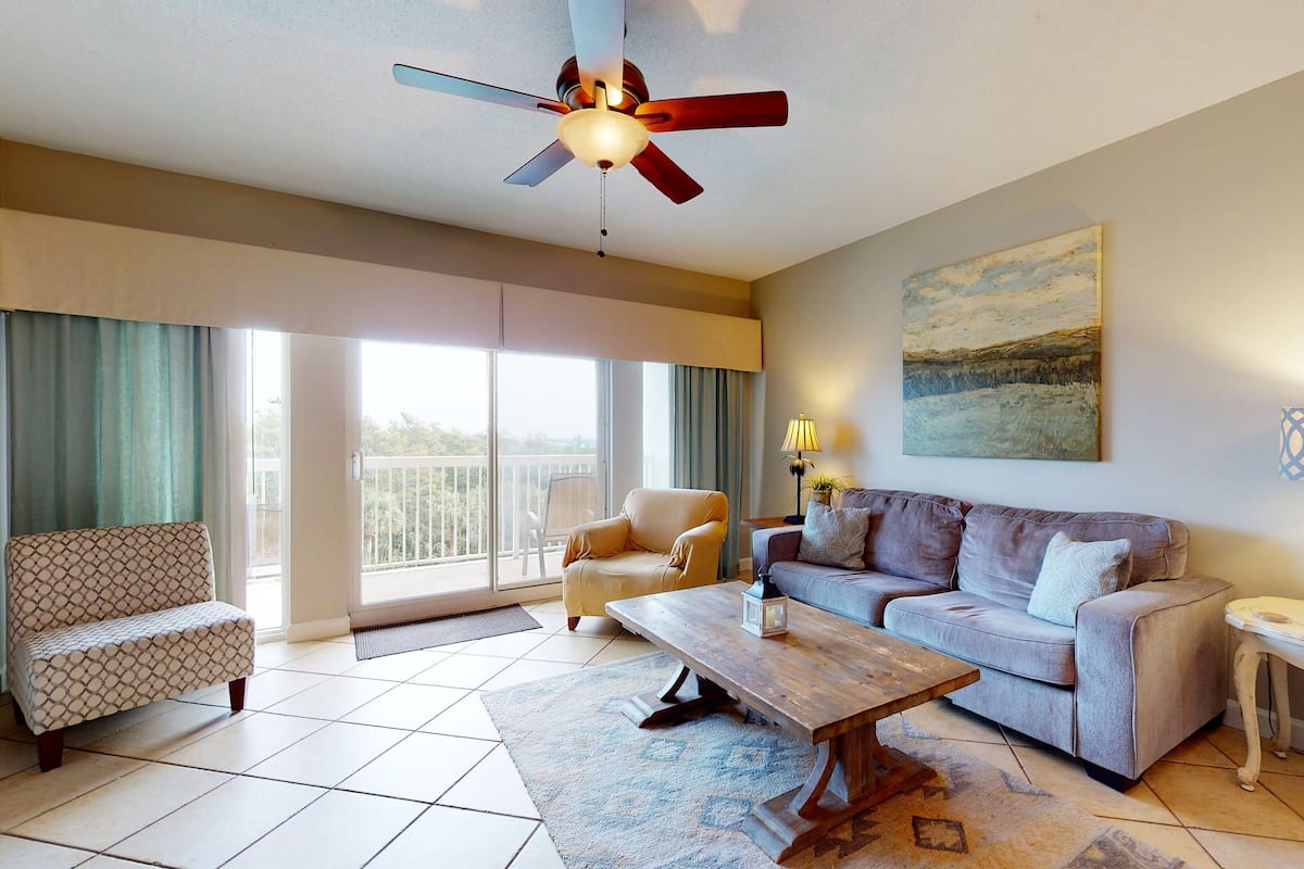 Ein großzügiges Wohnzimmer mit Sofa, Sessel und Couchtisch. An der Wand sind ein Deckenventilator und ein Gemälde zu sehen.