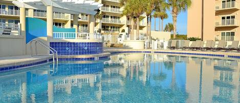 A swimming pool with a blue tiled edge, surrounded by a multi-story hotel with balconies and palm trees.