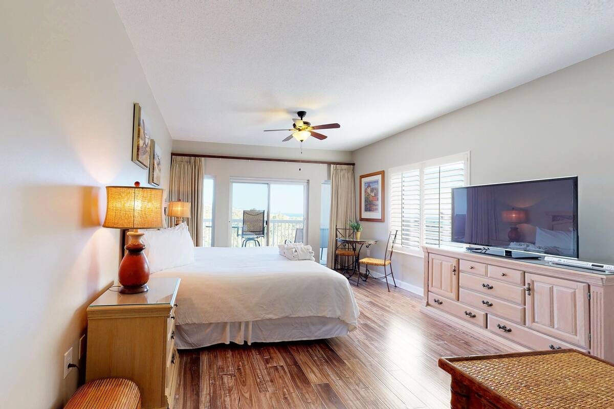 Ein Hotelzimmer mit Bett, Fernseher, Schreibtisch mit Lampe, Fenster mit Jalousien und Balkon mit Stuhl.