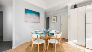 Un coin repas avec une table à plateau de verre, des chaises blanches et un tableau représentant une scène de plage.