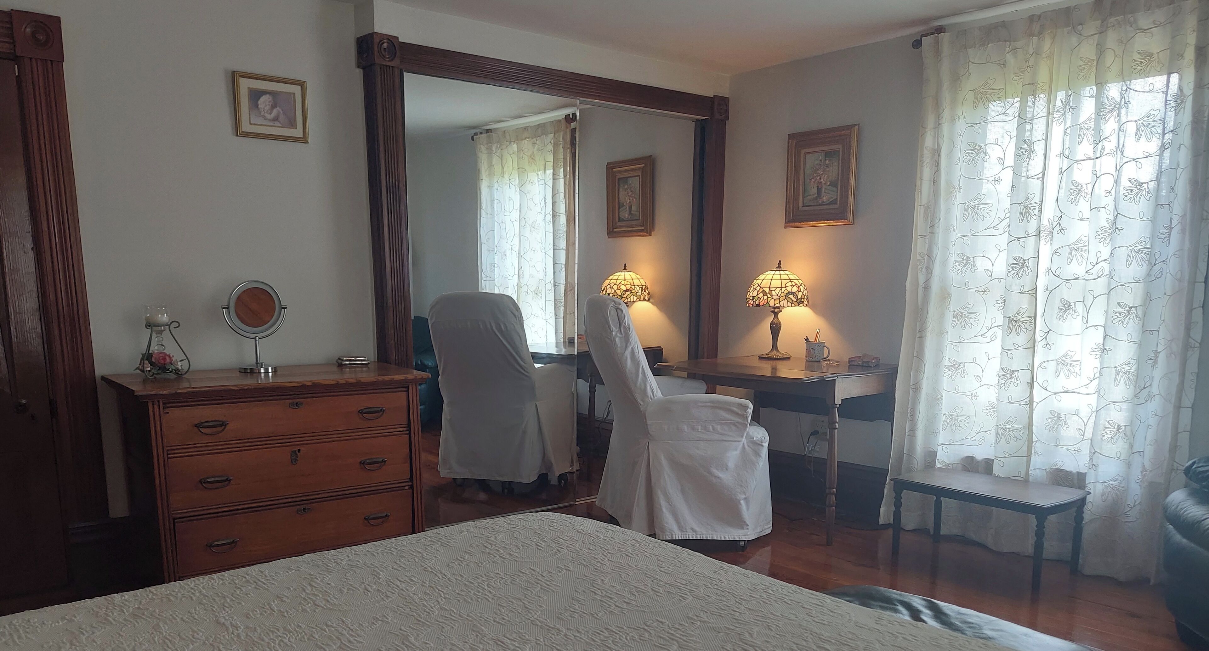 A bedroom with a bed, a dresser, a desk with a chair, a mirror, two lamps, and a window with curtains.