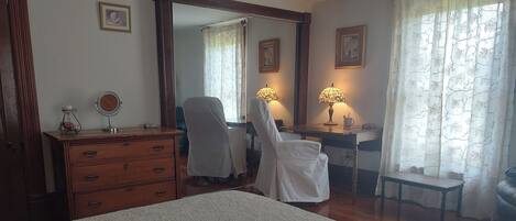 A bedroom with a bed, a dresser, a desk with a chair, a mirror, two lamps, and a window with curtains.