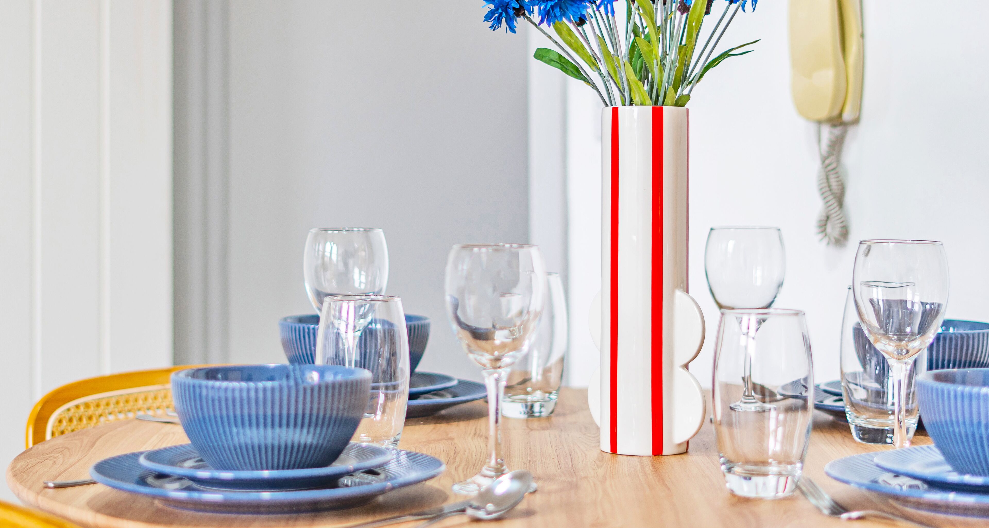 A dining table set with blue bowls, plates, and clear wine glasses, a striped vase with flowers, and a yellow telephone.