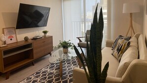 A modern living room with a flat-screen TV, a wooden entertainment unit, a glass coffee table, a beige sofa, and a large plant. - Redibuk - Premiere 1BR in San Isidro (Lima)