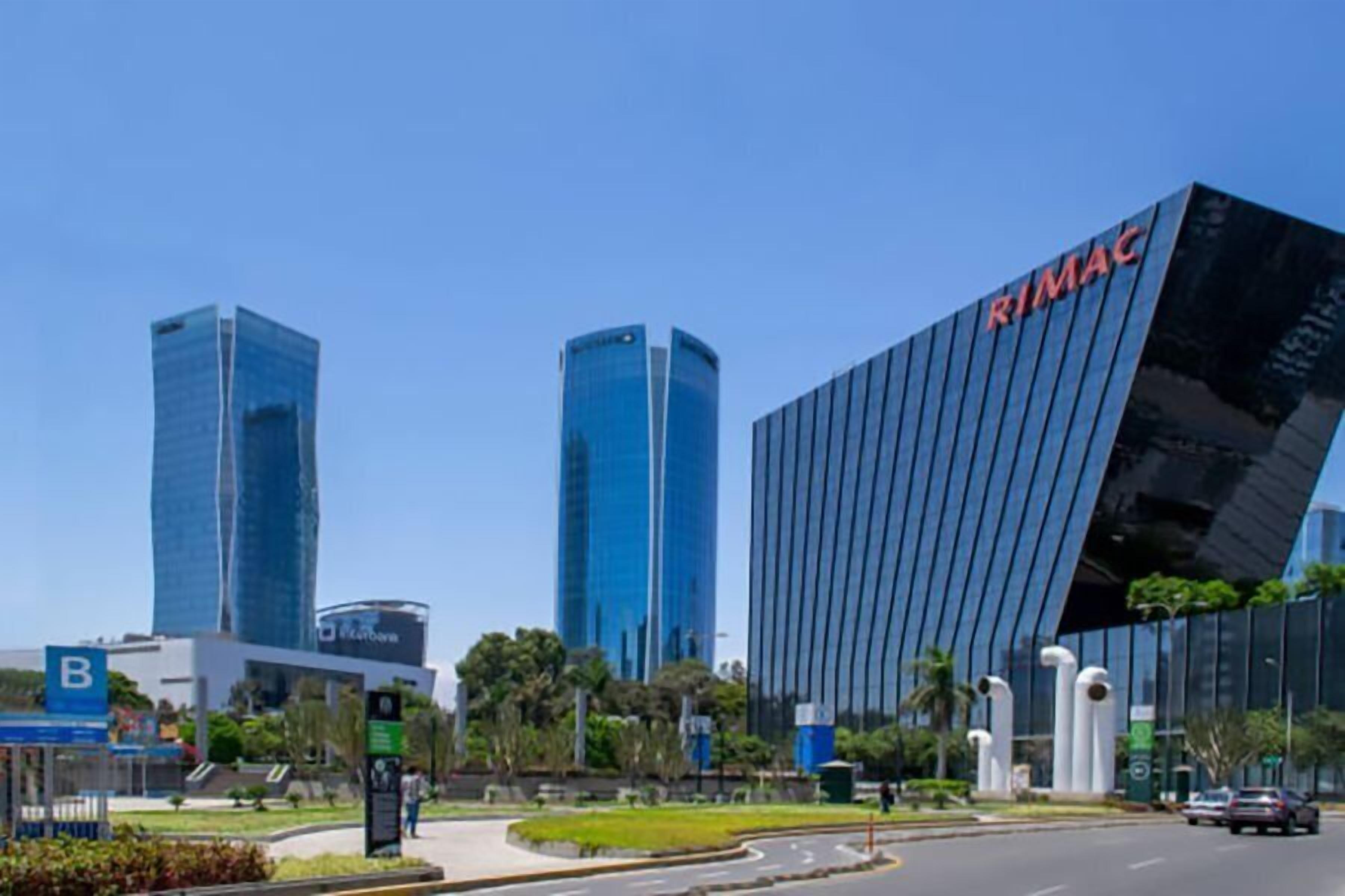Un edificio moderno con el letrero "RIMAC" en su fachada, rodeado de vegetación y un cielo azul despejado.