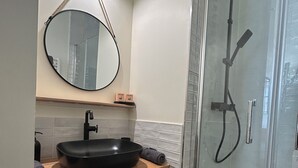 A bathroom with a round mirror, black sink, and a glass-enclosed shower. - Appartement Centre Ville (Castelnaudary)