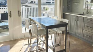 Une cuisine moderne avec une table à manger en verre et des chaises blanches, un balcon doté d’une balustrade blanche et des armoires grises.