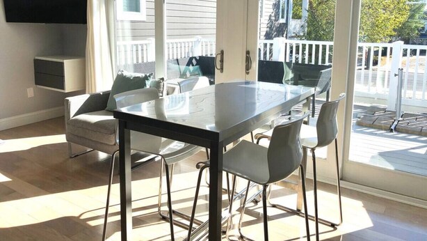 Un salon moderne avec une table à manger en verre, des sièges confortables, un téléviseur à écran plat fixé au mur et de grandes fenêtres donnant sur le jardin.