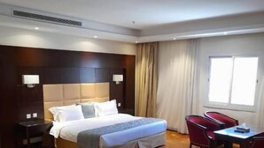 A hotel room with a large bed, a desk with a chair, a window, and a wall-mounted air conditioning unit.