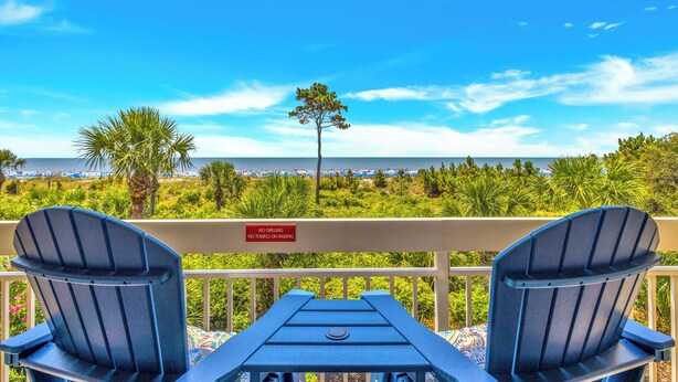 A balcony with two Adirondack chairs facing a beach. A sign on the railing reads "NO GRILLING NO TOWELS ON RAILING."