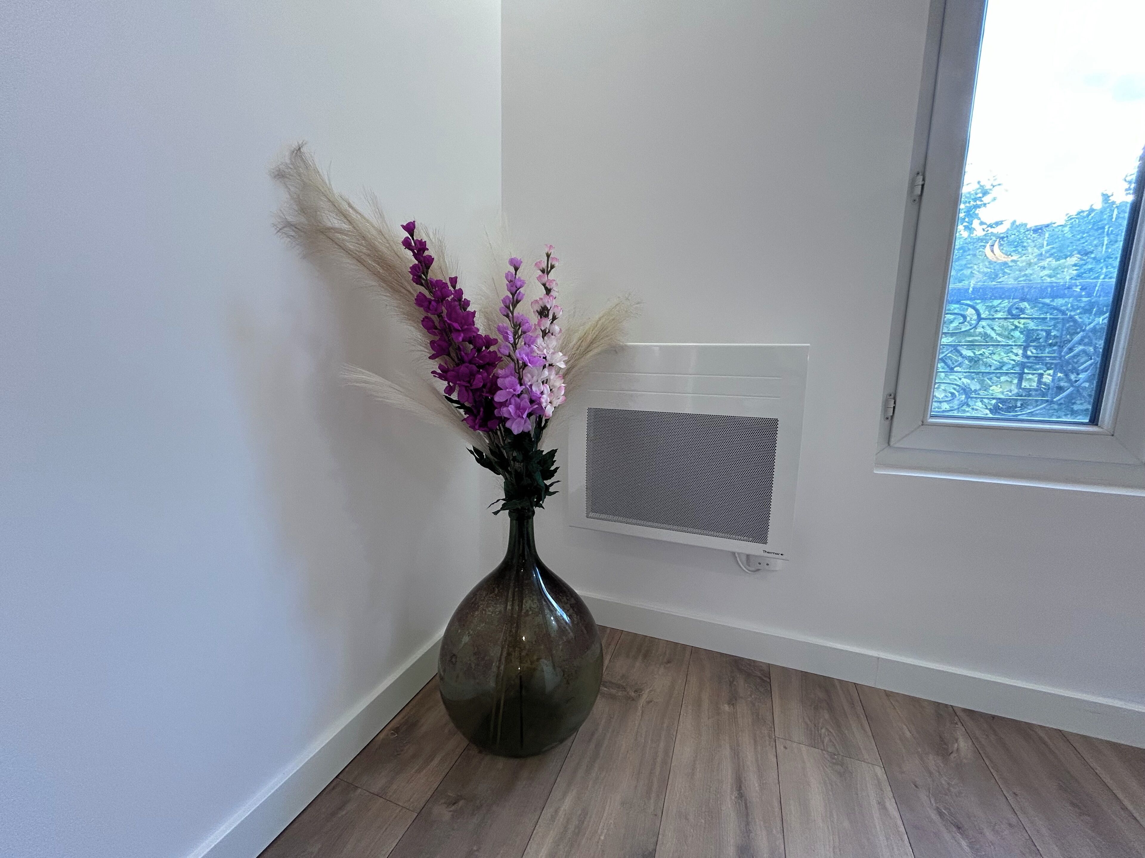 A room with a large window, a radiator, and a vase with flowers on the floor.