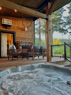 A wooden cabin with a hot tub, stone fireplace, and outdoor seating area with a view of trees and hills.